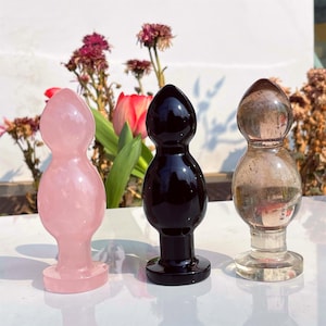 May include: Three decorative butt plugs in different colours. One is pale pink, one is black, and the third is clear. Each has a rounded top and a flared base. The objects are displayed on a white surface with flowers in the background.