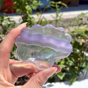 Op de afbeelding: Een doorschijnend, lichtpaars uitgesneden stenen object in de vorm van een wolk met een glad, gepolijst oppervlak. De steen heeft horizontale banden in verschillende tinten lavendel en heldere, kleurloze secties met een gelaagd effect.