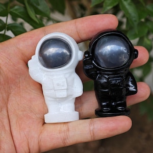 Könnte beinhalten: Zwei Astronautenfiguren, eine weiße und eine schwarze, werden gezeigt. Die Astronautenhelme haben ein reflektierendes, metallisches Aussehen. Der weiße Astronaut winkt, während der schwarze Astronaut den Arm hebt. Die Figuren sind klein und detailliert.