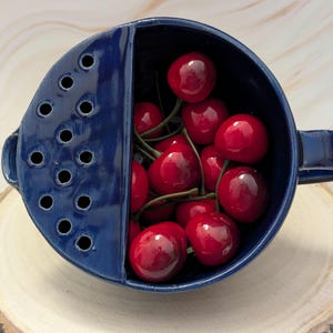May include: A deep blue ceramic berry bowl with a handle and a built-in colander section on one side, filled with a cluster of bright red cherries. The bowl features a smooth, glossy finish and a series of circular drainage holes.
