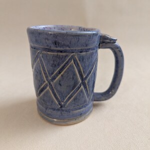 May include: A blue ceramic mug with a geometric diamond pattern carved into the exterior. The mug has a curved handle and a textured surface. The mug is approximately 4 inches tall.