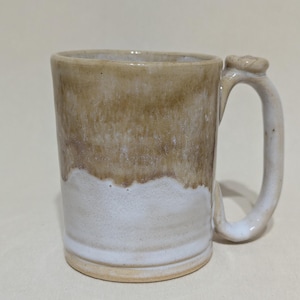 May include: A handmade ceramic mug with a unique handle design. The mug features a two-tone glaze, with a light brown upper section and a white lower section. The handle is a curved shape with a decorative element at the top. The mug is suitable for hot or cold beverages.