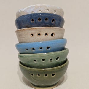 May include: A stack of handmade ceramic bowls in various colors, including white, blue, and green. Each bowl has a series of small holes along the rim. The bowls are stacked on top of each other, showcasing their different hues and textures.