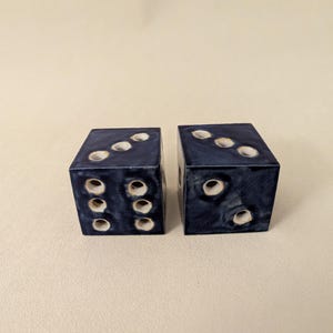 May include: Two navy blue dice with white dot markings. Each die has a cube shape with rounded edges. The dice are displayed on a neutral background. The dice are used for games.