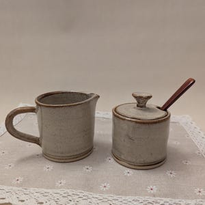May include: A ceramic creamer and sugar bowl set. The creamer has a handle and spout. The sugar bowl has a lid and a wooden spoon. Both pieces are light gray with brown trim and sit on a floral patterned placemat.