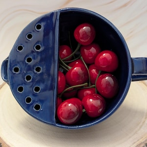 May include: A deep blue ceramic berry bowl with a built-in strainer section on one side, featuring a glossy finish and a handle on the right. The bowl is filled with a cluster of bright red, shiny cherries with green stems visible inside.