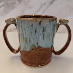 May include: A ceramic mug featuring two handles on opposite sides. The upper portion of the mug has a light blue glaze with dark brown vertical streaks, while the lower portion and handles are finished in a textured, earthy brown glaze.