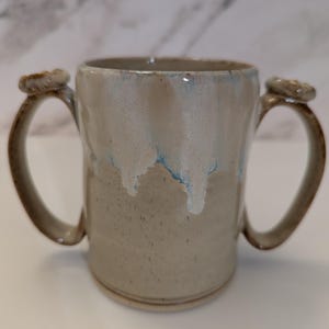May include: A ceramic mug featuring two side handles and a speckled beige base. The upper portion of the mug is coated in a glossy white glaze that drips down over the beige surface, revealing subtle hints of blue within the white glaze texture.