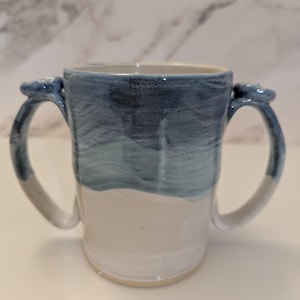 May include: A ceramic mug featuring two handles on opposite sides. The upper portion of the mug is glazed in a gradient of dark blue to light blue, while the lower portion remains a solid off-white. The handles are also glazed in blue.