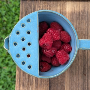 May include: A light blue ceramic berry bowl with a built-in strainer section on one side and a handle on the other. The bowl is filled with fresh, bright red raspberries. The exterior and interior surfaces feature a smooth, glossy finish.