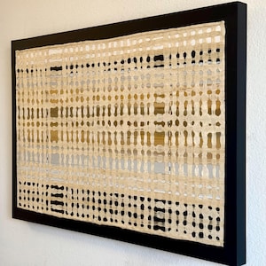 May include: Abstract artwork in a black frame. The piece features a textured, woven design in cream and tan hues, with a grid-like pattern. The artwork has a modern aesthetic and is mounted on a white wall.