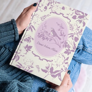Puede incluir: Un diario con diseño floral en crema y lavanda. La portada presenta un óvalo con un pájaro posado en una rama y el texto "Dear Mom Alice". El diario está sostenido en las manos de una persona, que lleva un jersey azul y vaqueros.