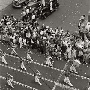 Puede incluir: Una fotografía en blanco y negro de una banda de música marchando en un desfile. La banda marcha por una calle llena de gente. La banda toca instrumentos y lleva banderas. Hay coches aparcados a un lado de la calle.