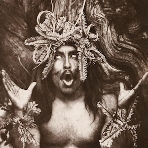 Puede incluir: Un hombre con el pelo largo y una corona de ramitas y ramas en la cabeza, con un collar de ramitas y ramas, está de pie frente a un tronco de árbol. Está mirando hacia arriba y tiene la boca abierta como si estuviera gritando. La imagen está en blanco y negro.