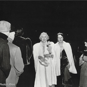 May include: Black and white photo of four people in formal attire. Two women are wearing white fur coats, one woman is wearing a black dress with a fur coat, and a man is wearing a dark suit and a hat.