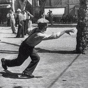 Puede incluir: Una fotografía en blanco y negro que muestra a una persona con gorra plana, camisa clara y pantalones oscuros con tirantes, captada en un movimiento dinámico hacia adelante mientras lanza una bola de metal en la petanca.