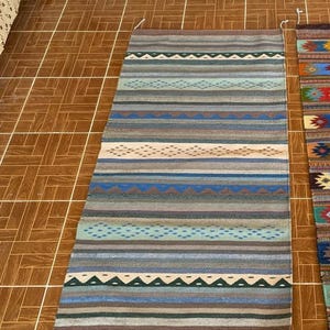 Könnte beinhalten: Ein rechteckiger, handgewebter Teppich mit einem Streifenmuster in Blau-, Braun- und Cremetönen. Der Teppich weist geometrische Muster auf und besteht wahrscheinlich aus Naturfasern. Der Teppich eignet sich für ein Wohn- oder Schlafzimmer.