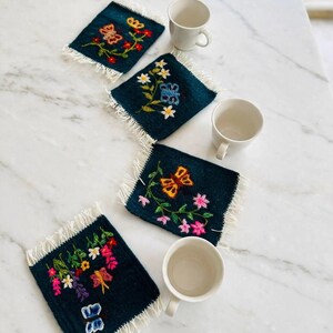Puede incluir: Cuatro posavasos rectangulares de tela azul oscuro con bordes de flecos blancos, cada uno con diseños únicos bordados a mano de mariposas coloridas y varios tallos florales, acompañados de tres tazas de cerámica blanca lisa.