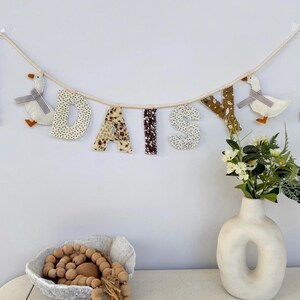 May include: A decorative banner spelling "DAISY" in fabric letters with various patterns. The banner is strung on a rope and features two goose ornaments. A white vase with flowers and a bowl with wooden beads are also in the image.