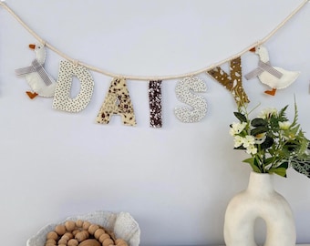May include: A decorative banner spelling "DAISY" in fabric letters with various patterns. The banner is strung on a rope and features two goose ornaments. A white vase with flowers and a bowl with wooden beads are also in the image.