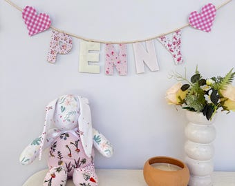 May include: A handmade bunny plush toy with floral fabric ears and a pink body with a garden vegetable print. A banner spelling "PENNY" hangs above, with gingham hearts and floral letters. A candle and vase are also on the table.