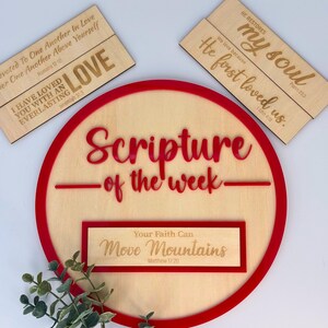 May include: A wooden round sign with a red border that says "Scripture of the week."  A smaller wooden sign with the text "Your Faith Can Move Mountains Matthew 17:20" is attached to the larger sign.