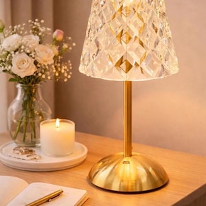 May include: A decorative table lamp with a faceted crystal shade and a gold-colored base and stem. The lamp is illuminated, casting a warm glow. The lamp is on a wooden surface, next to a candle and a notebook.