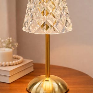 Può includere: Lampada da tavolo decorativa con paralume in vetro trasparente sfaccettato e base e stelo in metallo color oro. La lampada è accesa, emettendo un bagliore caldo. La lampada è su un tavolo di legno.