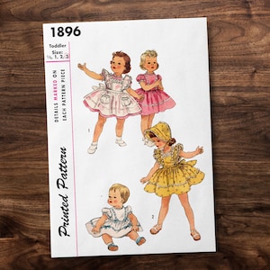 May include: Vintage sewing pattern for toddler dresses. The pattern is for sizes 1/2, 1, 2, and 3. The pattern shows four different dresses, one in pink, one in yellow, and two in white. The pattern is titled "1896" and "Printed Pattern".