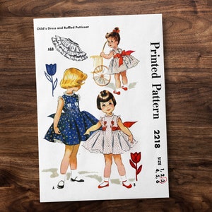 May include: Vintage sewing pattern for a child's dress and ruffled petticoat. The pattern is printed on white paper with a red and white floral design. The pattern number is 2218 and the sizes are 1, 2, 4, 5, and 6.