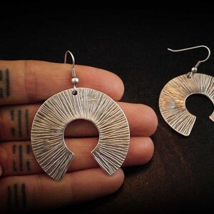 May include: A pair of silver earrings with a textured, sunburst design. The earrings are in the shape of a crescent moon with a cut-out circle in the centre.