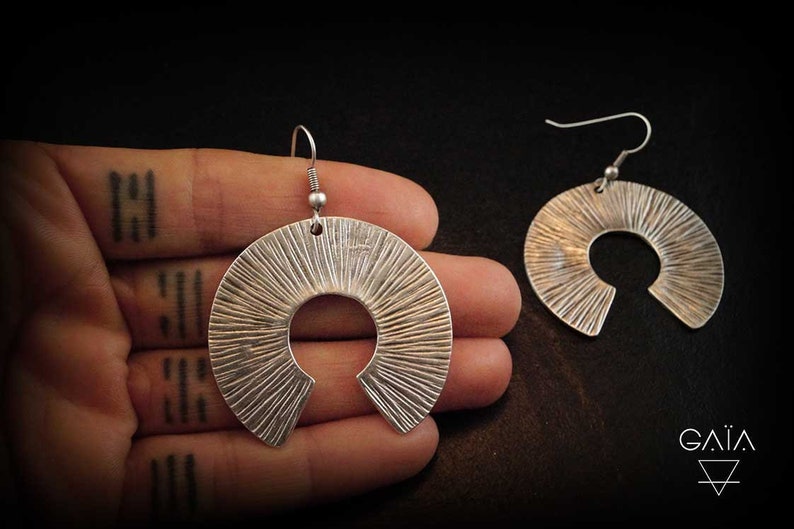 May include: A pair of silver earrings with a textured, sunburst design. The earrings are in the shape of a crescent moon with a cut-out circle in the centre.