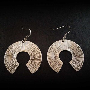 May include: A pair of silver hoop earrings with a textured, sunburst design. The earrings have a large, open circle shape.