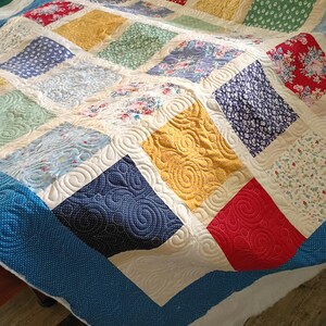 May include: A patchwork quilt featuring a grid of multi-colored fabric squares in shades of red, yellow, blue, and green with floral and geometric patterns. The quilt is finished with a cream-colored border and intricate swirl quilting.