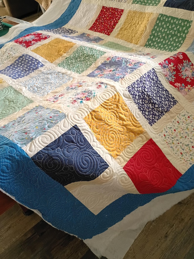 May include: A patchwork quilt featuring a grid of multi-colored fabric squares in shades of red, yellow, blue, and green with floral and geometric patterns. The quilt is finished with a cream-colored border and intricate swirl quilting.