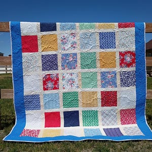 May include: A patchwork quilt featuring a grid of square fabric blocks in various colors and patterns, including solid red, yellow, blue, and green, alongside floral and plaid prints. The quilt has a bright blue border and white stitching.