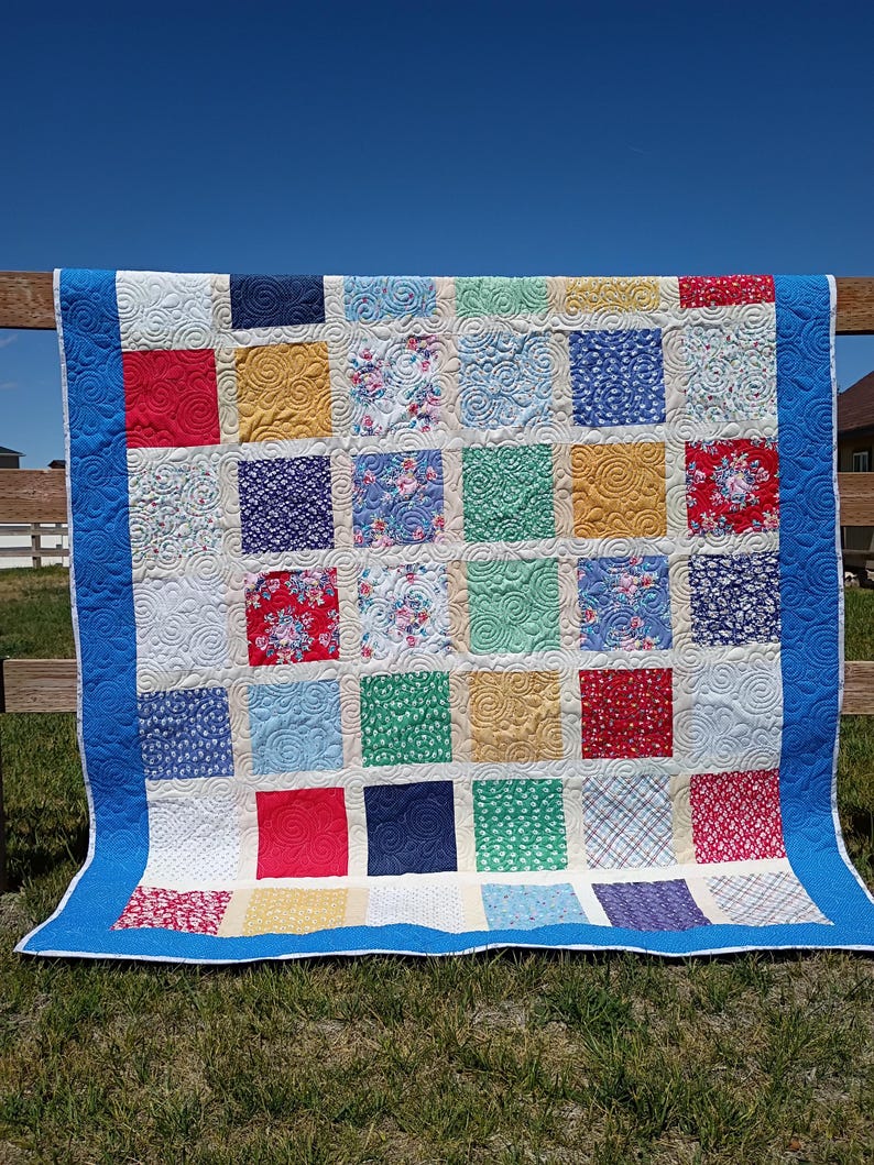 May include: A patchwork quilt featuring a grid of square fabric blocks in various colors and patterns, including solid red, yellow, blue, and green, alongside floral and plaid prints. The quilt has a bright blue border and white stitching.