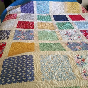 May include: A patchwork quilt featuring a grid of multi-colored fabric squares in shades of blue, yellow, red, green, and white. The quilt displays various floral and geometric patterns, all unified by a cream-colored sashing with a swirling stitch.