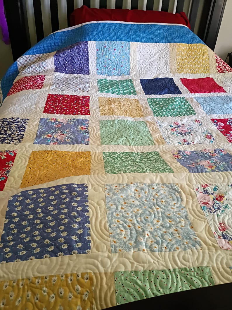 May include: A patchwork quilt featuring a grid of multi-colored fabric squares in shades of blue, yellow, red, green, and white. The quilt displays various floral and geometric patterns, all unified by a cream-colored sashing with a swirling stitch.