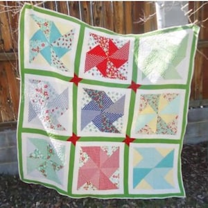 Peut inclure: Une courtepointe en patchwork avec un motif de moulin à vent. La courtepointe est faite de différents tissus dans des tons de rouge, bleu, vert et blanc. La courtepointe est suspendue à une corde à linge.