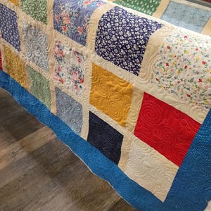 May include: A patchwork quilt featuring a grid of multi-colored fabric squares in shades of red, blue, yellow, and green, separated by cream-colored sashing. The quilt displays intricate swirling machine-quilted patterns across the surface.