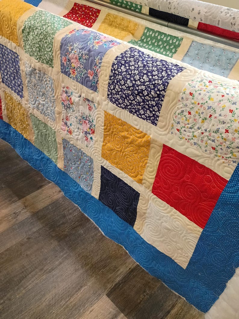 May include: A patchwork quilt featuring a grid of multi-colored fabric squares in shades of red, blue, yellow, and green, separated by cream-colored sashing. The quilt displays intricate swirling machine-quilted patterns across the surface.
