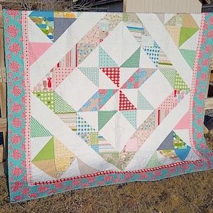 Op de afbeelding: Een handgemaakte quilt met een witte achtergrond en kleurrijke geometrische patronen. De quilt heeft driehoeken in verschillende kleuren, waaronder rood, groen, roze en blauw. Een turquoise rand met een rood bloemenpatroon omlijst de quilt.