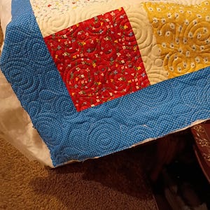 May include: A patchwork quilt featuring square fabric blocks in red, yellow, blue, and white with floral patterns. The quilt is finished with a blue border featuring a white polka dot pattern and is stitched with a continuous swirling pattern.