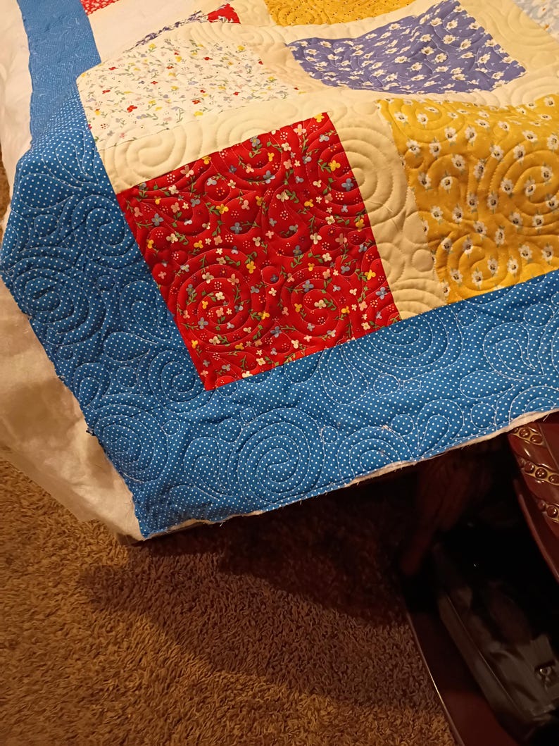 May include: A patchwork quilt featuring square fabric blocks in red, yellow, blue, and white with floral patterns. The quilt is finished with a blue border featuring a white polka dot pattern and is stitched with a continuous swirling pattern.