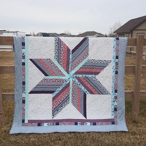 May include: A quilt featuring a large central eight-pointed star made of strips in shades of red, white, blue, and grey. The background consists of light grey fabric with a circular fan quilting pattern, framed by a border of small patterned squares.