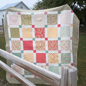 May include: A patchwork quilt featuring a grid of square fabric blocks in shades of red, green, cream, and yellow. The quilt has a wide olive green border with a small-scale pattern and is constructed with small solid-colored corner squares.