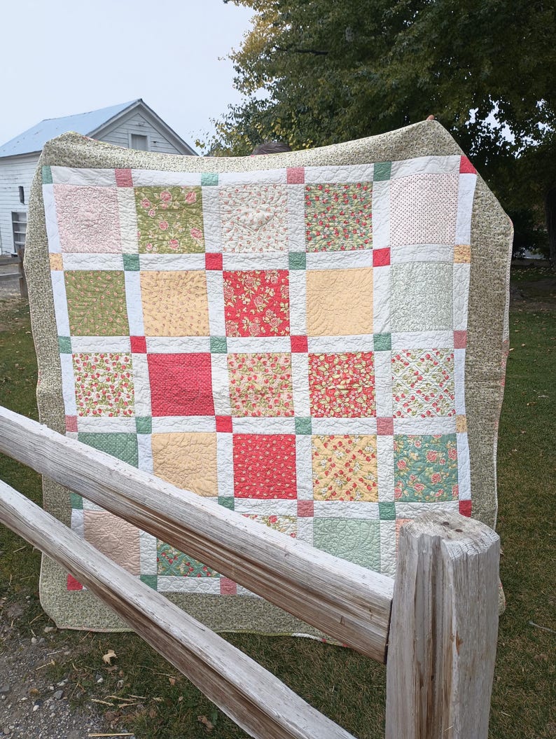 May include: A patchwork quilt featuring a grid of square fabric blocks in shades of red, green, cream, and yellow. The quilt has a wide olive green border with a small-scale pattern and is constructed with small solid-colored corner squares.