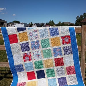 May include: A patchwork quilt featuring a grid of multi-colored fabric squares in shades of red, blue, yellow, green, and white. The quilt is bordered by a solid blue fabric and displays a continuous swirling quilted pattern across the surface.