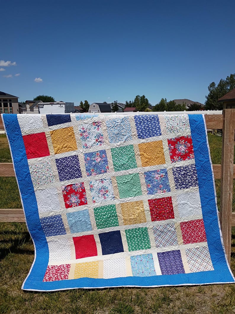 May include: A patchwork quilt featuring a grid of multi-colored fabric squares in shades of red, blue, yellow, green, and white. The quilt is bordered by a solid blue fabric and displays a continuous swirling quilted pattern across the surface.